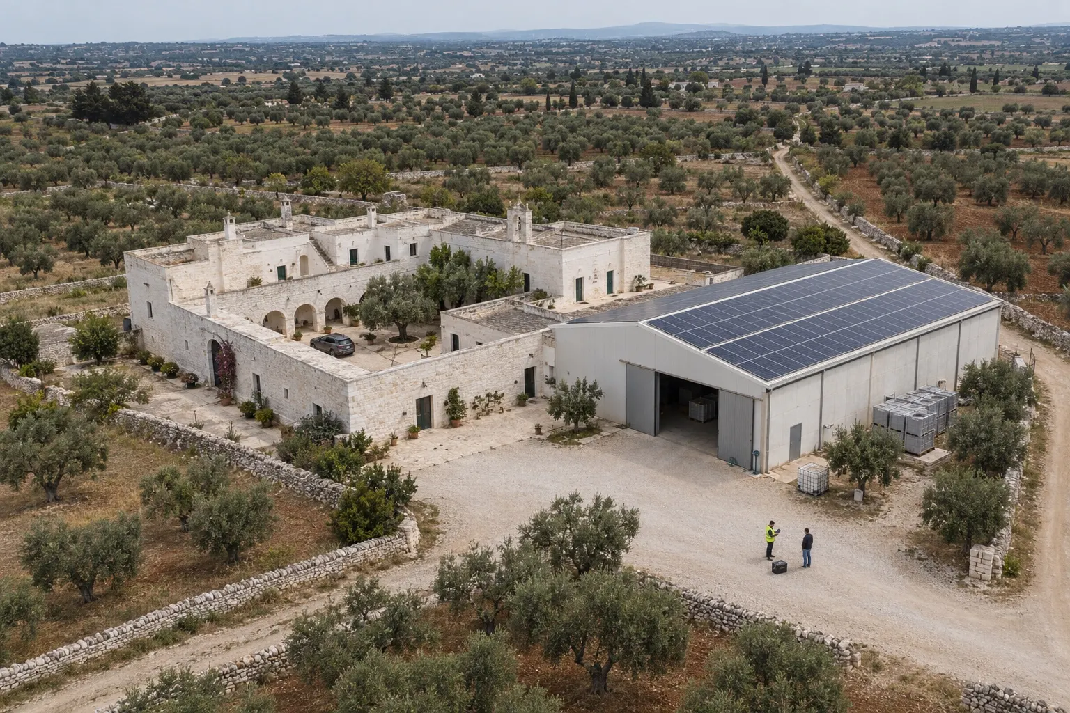 Vista aerea di una masseria pugliese in pietra bianca con corte interna e annesso capannone agricolo con impianto fotovoltaico in copertura, circondata da uliveti e muretti a secco; nel piazzale un tecnico con giubbotto ad alta visibilità e tablet parla con il committente