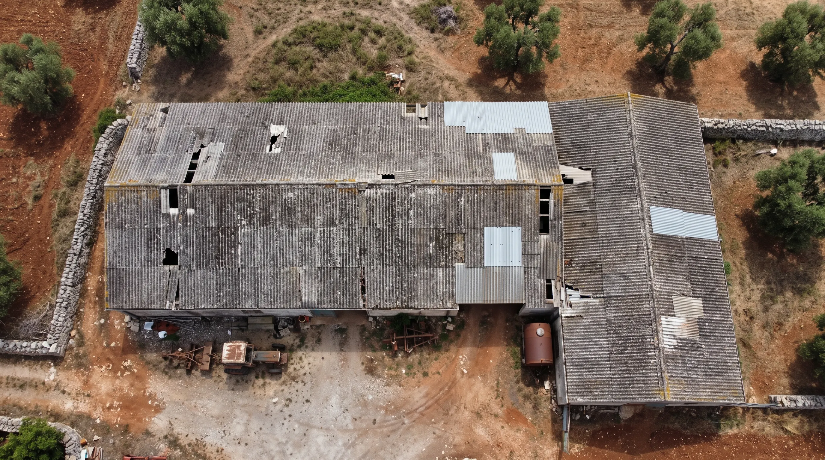 Vista zenitale da drone di capannoni agricoli con coperture in lastre ondulate di fibrocemento annerite da licheni, lastre rotte o mancanti e alcune sostituzioni in lamiera; nel piazzale un vecchio trattore e una cisterna arrugginita, attorno terra rossa, ulivi e muretti a secco